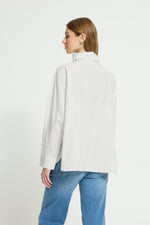 Blusa blanca con raya diplomática fina en marrón, cuello alto vuelto y botones decorativos. Un diseño elegante y versátil para tus looks de temporada.